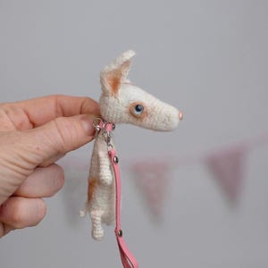 May include: A handmade, crocheted dog figurine with a pink collar and leash. The dog is white with tan markings and blue eyes. The leash is a light pink color. The dog is held by a person's hand.