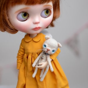 May include: A Blythe doll with auburn hair and green eyes wearing a mustard yellow dress. The doll is holding a small, white crocheted cat with blue eyes and a silver heart pendant. The doll's dress has a Peter Pan collar.