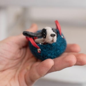 May include: A handmade felted dog figurine with black and white markings peeks out of a blue felt carrier with red straps. The miniature dog is held in a person's open hand, showcasing the craft's detail.