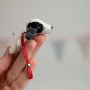 May include: A small, handmade white and black puppy figurine with a red leash. The puppy has a black patch over one eye and floppy ears. The leash is attached with a silver button.