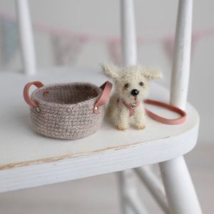 May include: A miniature crochet basket with pink leather handles sits next to a small, fluffy white dog figurine. The dog wears a pink collar and has a matching leash. The items are on a white chair.