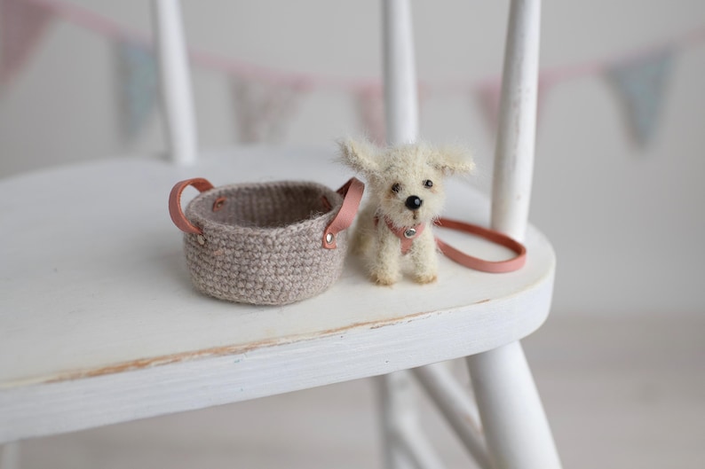 May include: A miniature crochet basket with pink leather handles sits next to a small, fluffy white dog figurine. The dog wears a pink collar and has a matching leash. The items are on a white chair.