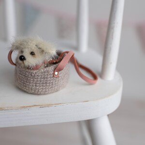May include: A miniature plush dog peeks out of a small, crocheted basket with a pink leather handle. The basket is a neutral beige color, and the dog has fluffy, light-colored fur. The scene is set on a white wooden chair.