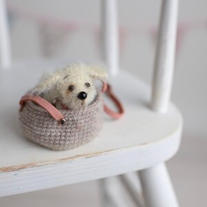 May include: A small, plush, cream-colored dog toy peeks out of a crocheted, taupe-colored basket with a pink leather handle. The toy has black eyes and a black nose. The basket is on a white wooden chair.