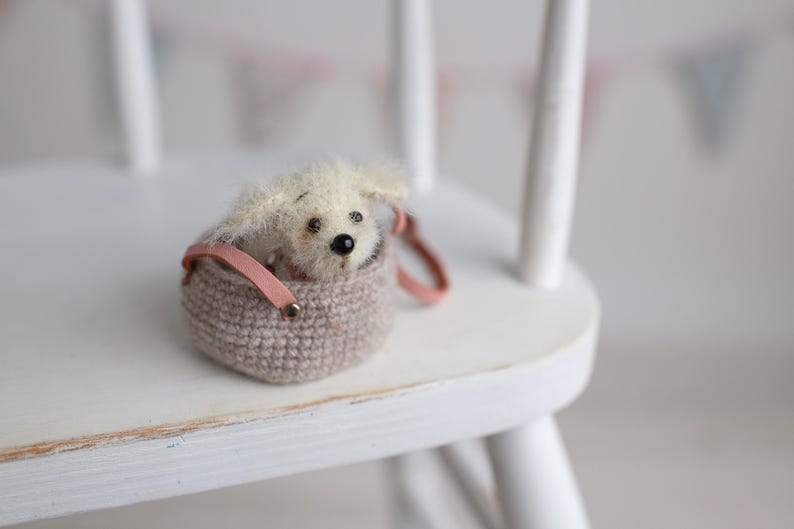 May include: A small, plush, cream-colored dog toy peeks out of a crocheted, taupe-colored basket with a pink leather handle. The toy has black eyes and a black nose. The basket is on a white wooden chair.