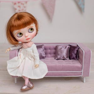 May include: A doll with red hair and a white dress stands next to a miniature pink sofa with three purple velvet pillows. The doll is wearing pink shoes and a pink collar. Decorative bunting hangs in the background.