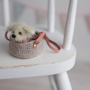 May include: A miniature plush dog peeks out of a small, crocheted basket with a pink leather handle. The basket is a neutral beige color, and the dog has fluffy, light-colored fur. The scene is set on a white wooden chair.