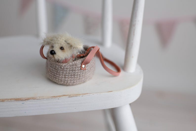 May include: A miniature plush dog peeks out of a small, crocheted basket with a pink leather handle. The basket is a neutral beige color, and the dog has fluffy, light-colored fur. The scene is set on a white wooden chair.