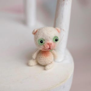 May include: A small, handcrafted needle-felted kitten figurine with off-white wool fur, large translucent green glass eyes, a soft pink nose, and delicate white whiskers. The miniature cat has a rounded body and sits in a seated position.