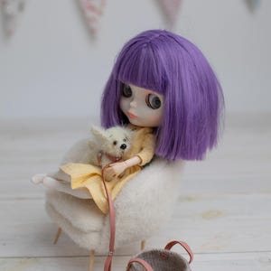 May include: A doll with purple hair and a yellow dress sits in a white chair, holding a small white dog. A small, gray woven basket with pink handles sits nearby. A pink banner hangs in the background.