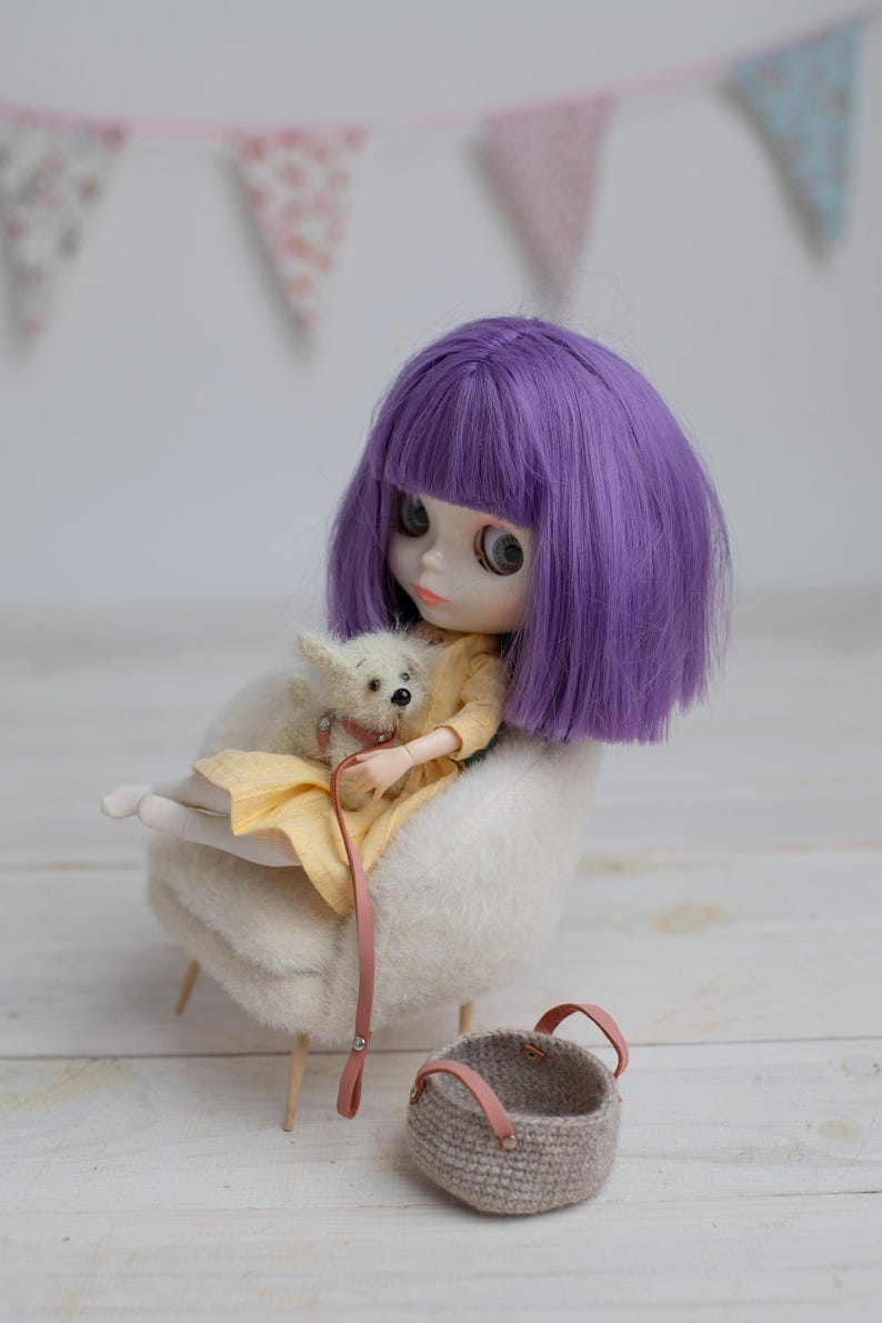 May include: A doll with purple hair and a yellow dress sits in a white chair, holding a small white dog. A small, gray woven basket with pink handles sits nearby. A pink banner hangs in the background.