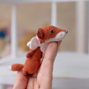 Op de afbeelding: Een klein gehaakt vosje met oranje en witte vacht, dat een roze vleugel vasthoudt. De vos heeft zwarte ogen en een witte snuit.