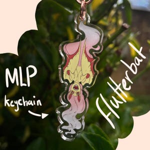 May include: A clear acrylic keychain with a cartoon bat design in pink, yellow, and white. The keychain hangs from a copper-coloured ring. The image includes the text "MLP keychain" and "Flutterbat".