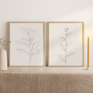 May include: Two minimalist line art prints of plants in black and white. The prints are framed in light wood and are displayed on a light brown linen tablecloth.