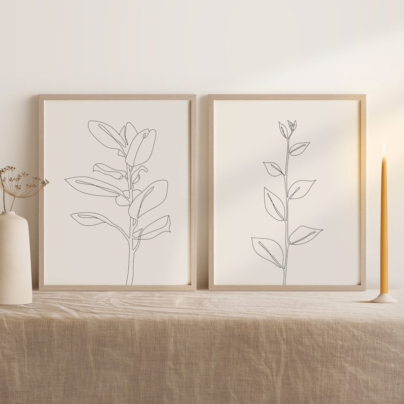 May include: Two minimalist line art prints of plants in black and white. The prints are framed in light wood and are displayed on a light brown linen tablecloth.