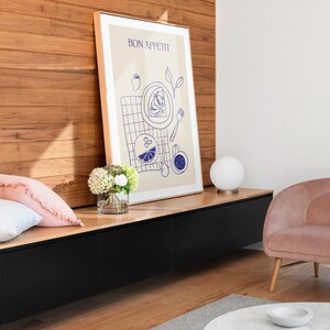 May include: A framed print with the text "BON APPETIT" in a blue font. The print features a blue illustration of a plate of food, a fork, a glass of water, and a cup of coffee. The print is hanging on a wall with a wooden paneling background.