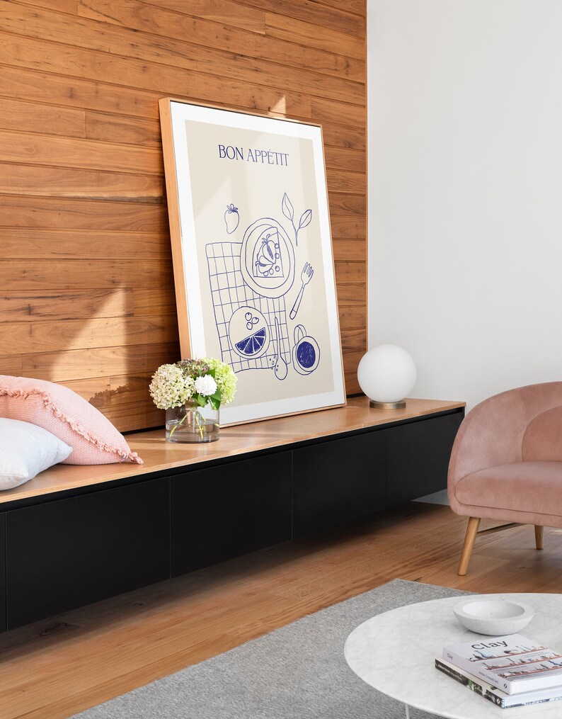 May include: A framed print with the text "BON APPETIT" in a blue font. The print features a blue illustration of a plate of food, a fork, a glass of water, and a cup of coffee. The print is hanging on a wall with a wooden paneling background.