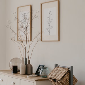 May include: A white dresser with three drawers and two doors, decorated with dried branches in three ceramic vases, a glass globe, a candle, a framed photo, and a glass jar.  Two framed prints with black line drawings of plants hang on the wall above the dresser. A gray metal stand with three wicker baskets sits on the right side of the dresser.