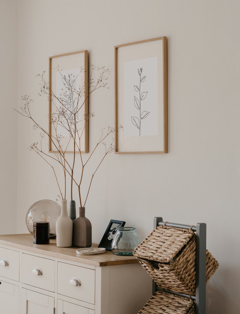 May include: A white dresser with three drawers and two doors, decorated with dried branches in three ceramic vases, a glass globe, a candle, a framed photo, and a glass jar.  Two framed prints with black line drawings of plants hang on the wall above the dresser. A gray metal stand with three wicker baskets sits on the right side of the dresser.