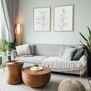 May include: A gray sectional sofa with pillows and a throw blanket sits in a living room with two wooden coffee tables, a gray rug, and two framed line drawings of plants on the wall.