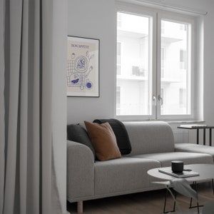 May include: A gray couch with brown and black throw pillows in a living room with a window and a framed print that says "Bon Appetit" in blue lettering. The print features a blue and white illustration of food and a table setting.