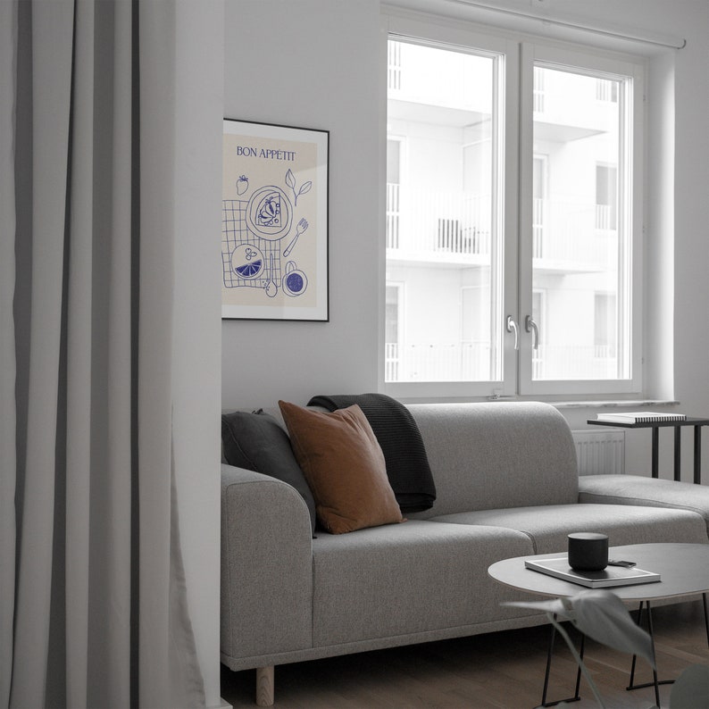 May include: A gray couch with brown and black throw pillows in a living room with a window and a framed print that says "Bon Appetit" in blue lettering. The print features a blue and white illustration of food and a table setting.