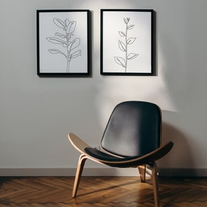 May include: Two black framed line drawings of plants hang on a white wall above a black leather chair with wooden legs.