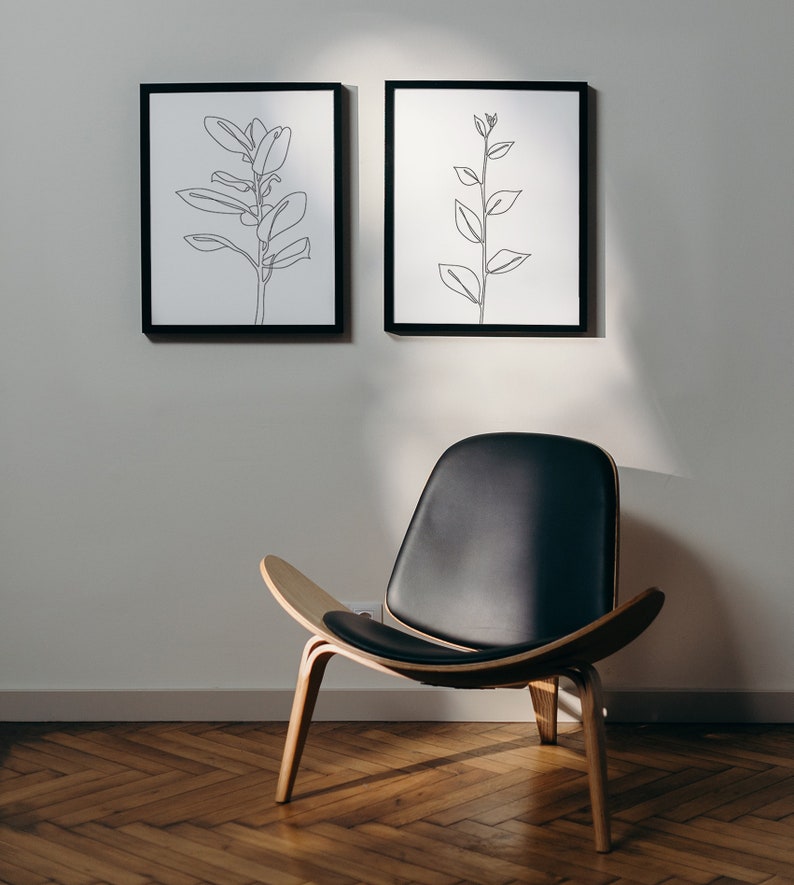 May include: Two black framed line drawings of plants hang on a white wall above a black leather chair with wooden legs.