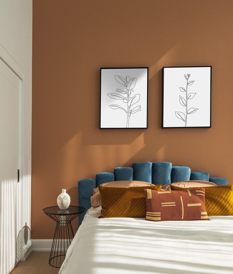 May include: A teal blue velvet headboard with a scalloped design. The headboard is decorated with a brown and gold patterned pillow and a white pillow with a brown geometric design. Two black framed prints with line drawings of plants hang on the wall behind the bed.