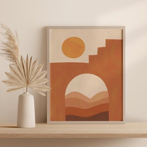 Puede incluir: Impresión de arte abstracto con un sol, un arco y colinas en capas en tonos de naranja, marrón y beige. La impresión está enmarcada en un marco de madera claro.