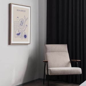 May include: A framed print with a beige background and blue illustrations of food and the text "BON APPETIT". The print is hanging on a light gray wall next to a beige armchair with black legs and wooden armrests.