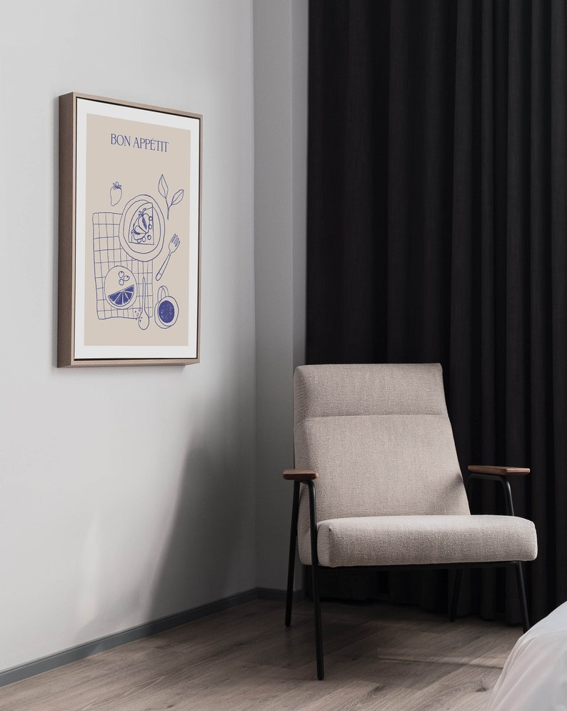 May include: A framed print with a beige background and blue illustrations of food and the text "BON APPETIT". The print is hanging on a light gray wall next to a beige armchair with black legs and wooden armrests.