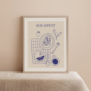 May include: A framed print with a beige background and blue line drawings of a plate of fruit, a glass of juice, a fork, a spoon, and a cup. The text "BON APP&Eacute;TIT" is at the top of the print.