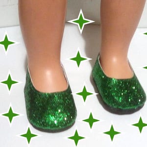 May include: A pair of green glitter doll shoes. The shoes are a flat style with a rounded toe and a low vamp. The glitter covers the entire shoe, giving it a sparkly appearance. The shoes are on a white surface with green star decorations.