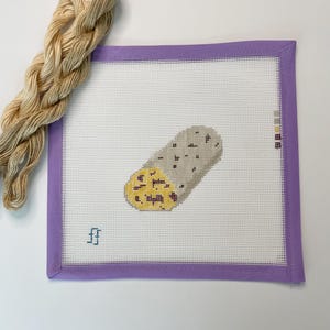May include: A needlepoint canvas featuring a burrito design, with a light purple border. A skein of beige embroidery floss is positioned to the left. The burrito is gray with yellow and black details. The canvas has a white mesh background.