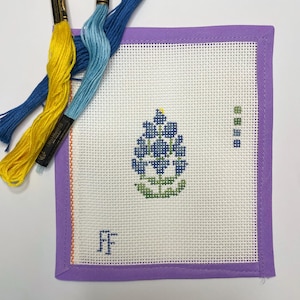 May include: A cross-stitch project featuring a bluebonnet design, framed in purple. Three skeins of embroidery floss in yellow, light blue, and dark blue are arranged near the project. The initials "FF" are stitched in blue in the lower left corner.
