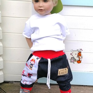 May include: A young child wearing a white t-shirt, a gray baseball cap with a green brim, red and blue denim pants with a fire truck design, and gray sneakers. The pants have a patch that says "klezmer rabauke".