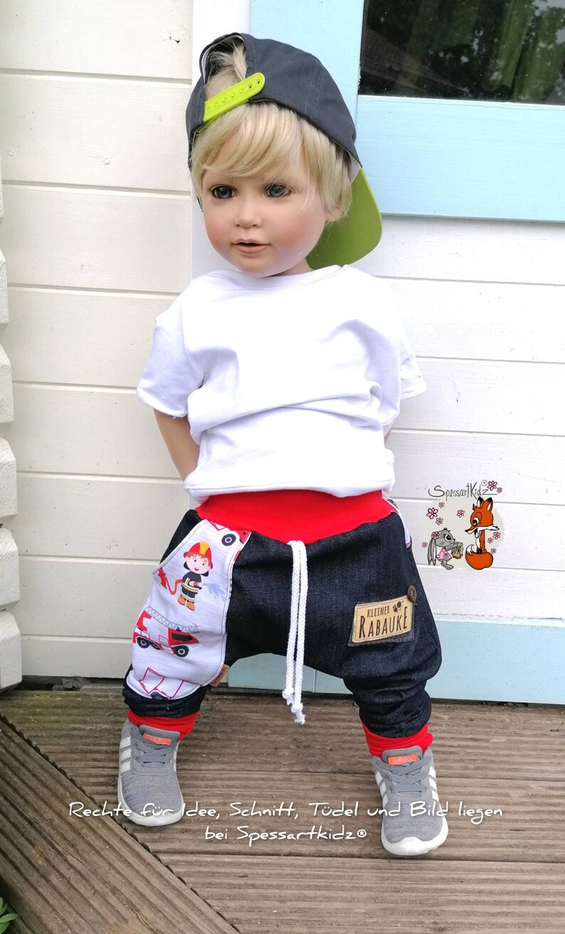 May include: A young child wearing a white t-shirt, a gray baseball cap with a green brim, red and blue denim pants with a fire truck design, and gray sneakers. The pants have a patch that says "klezmer rabauke".