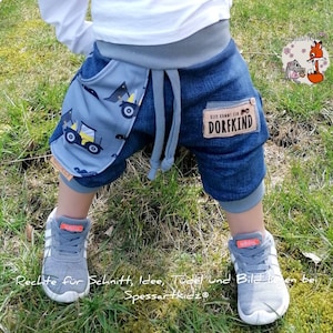 May include: A toddler wearing blue denim shorts with a gray and blue fabric pocket featuring a tractor design. The shorts have a gray waistband and a brown patch with the text "Hier kommt ein Dorfkind". The toddler is wearing gray sneakers with white laces.