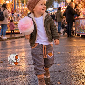 May include: A child wearing a brown knit beanie, a white t-shirt, an olive green hooded jacket with a cream-colored lining, and gray corduroy pants with a fox graphic on the pocket, holding a pink and white cotton candy treat.