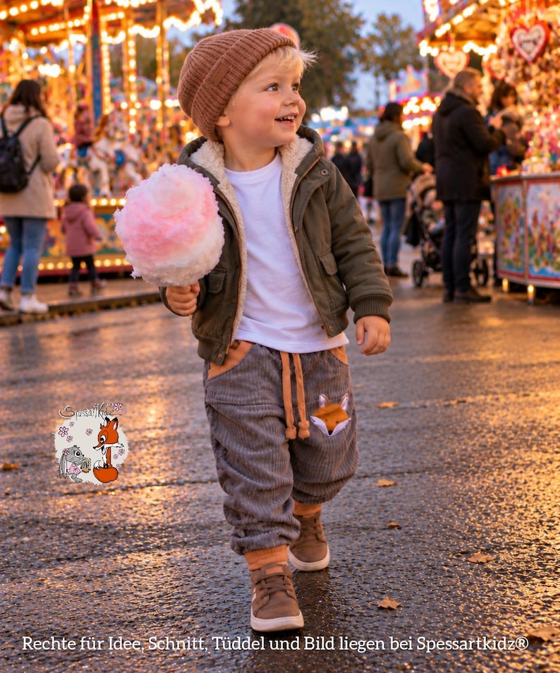 May include: A child wearing a brown knit beanie, a white t-shirt, an olive green hooded jacket with a cream-colored lining, and gray corduroy pants with a fox graphic on the pocket, holding a pink and white cotton candy treat.