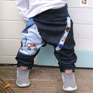 May include: A toddler wearing blue denim pants with a construction vehicle print on one leg. The pants have a black waistband and a button on the side. The toddler is wearing gray sneakers with orange accents.