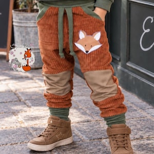 Könnte beinhalten: Rostfarbene Cordhose mit einem Fuchsmotiv als Applikation auf der linken Tasche, olivgrünen Rippbündchen und einem passenden olivgrünen Kordelzug am Bund. Die Hose ist mit hellbraunen Knieaufnähern versehen und wird mit braunen Sneakern kombiniert.