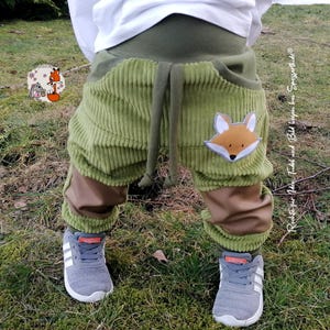 May include: Olive green corduroy pants featuring a wide elastic waistband, a drawstring, and a fox applique on the left leg. The pants have brown fabric panels at the lower legs and are paired with grey athletic sneakers and a white shirt.