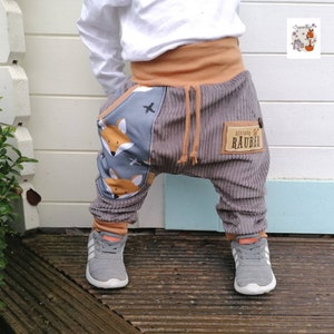 Könnte beinhalten: Graue Cordhose mit einem Fuchs-Druck an der Seite und einem Patch mit der Aufschrift "Kleiner Rauber". Die Hose hat einen braunen Gummizugbund und wird von einem Kind getragen.