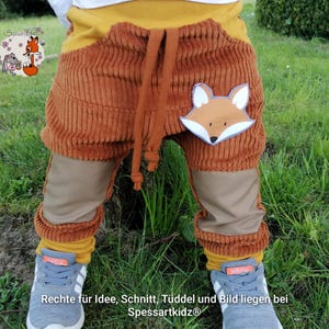 May include: Corduroy pants in shades of brown, mustard yellow, and tan, featuring a fox applique. The pants have a drawstring waist and are paired with gray sneakers. The text on the pants reads: "Rechte für Idee, Schnitt, Tüddel und Bild liegen bei Spessartkidz®."