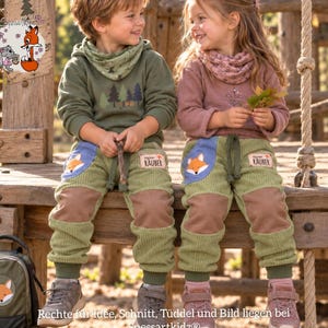 May include: Two children wearing olive green and dusty rose long-sleeve shirts with matching neck warmers and light green corduroy pants. The pants feature blue pockets with fox illustrations and patches labeled Kleiner R&auml;uber.