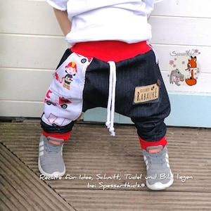 Könnte beinhalten: Ein Kind trägt eine blaue Jeanshose mit rotem Besatz und ein weißes Shirt. Die Hose hat auf einem Bein einen weißen und roten Feuerwehrwagen-Druck und auf dem anderen Bein einen blauen Patch mit dem Text "kleiner RAUAKE". Das Kind trägt graue Turnschuhe.