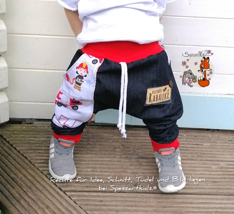 May include: A child wearing blue denim pants with red trim and a white shirt. The pants have a white and red fire truck print on one leg and a blue patch with the text "kleiner RAUAKE" on the other leg. The child is wearing gray sneakers.