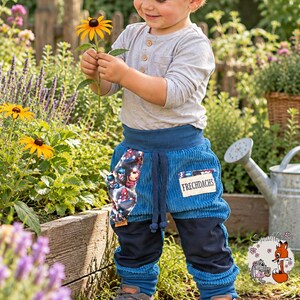 May include: A light gray long-sleeve henley shirt paired with blue corduroy pants featuring a superhero print pocket, a patch labeled FRECHDACHS, dark blue knee panels, and ribbed cuffs. The pants have a drawstring waist and a relaxed fit.